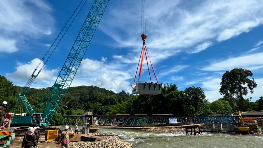 A precast UHPC beam segment is being hoisted by a crawler crane and transported under controlled lifting operations to the launching truss.